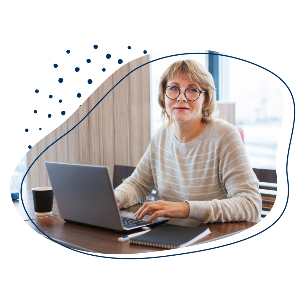 Altere Frau mit Brille schaut von ihrem Laptop hoch und lächelt in die Kamera.