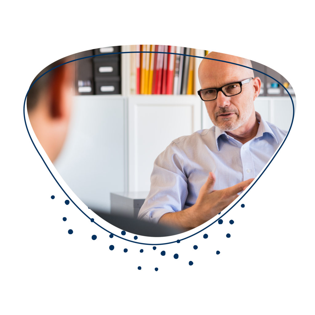 Mann mit Brille sitzt vor einem Regal und gestikuliert mit der Hand. Im Vordergrund ist eine weitere Person, jedoch unscharf zu erkennen.