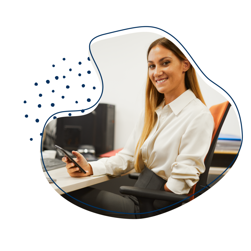 Frau Mit nur einem Arm sitzt an einem PC-Arbeitsplatz und hält ein Handy, dabei lächelt sie in die Kamera.