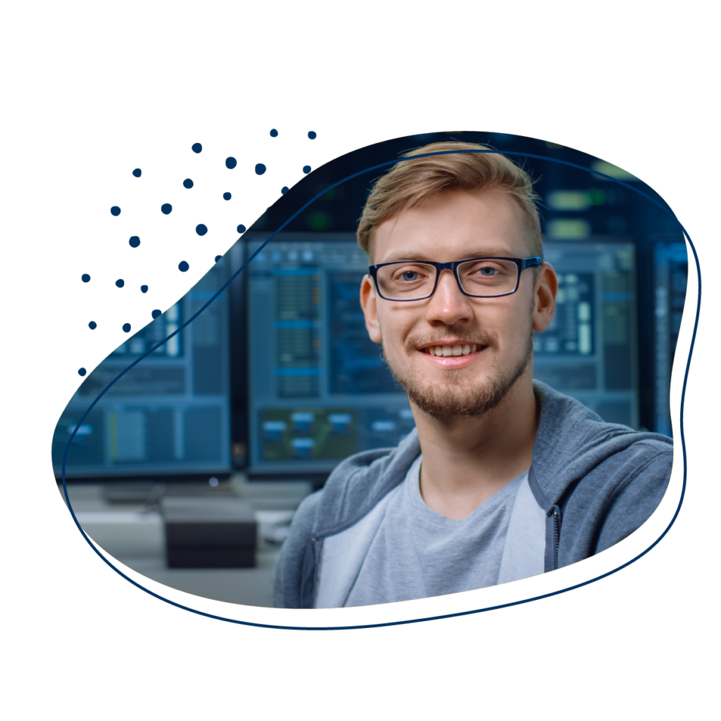 Lächelnder junger Mann mit Brille, der vor mehreren Computermonitoren sitzt.