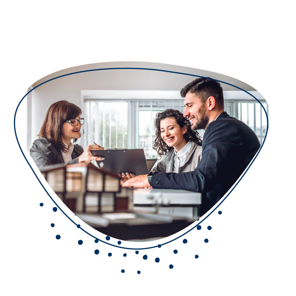 Frau sitzt mit einem Pärchen an einem Tisch und zeigt etwas in einem Dokument. m Vordergrund ist ein Miniaturhaus zu sehen