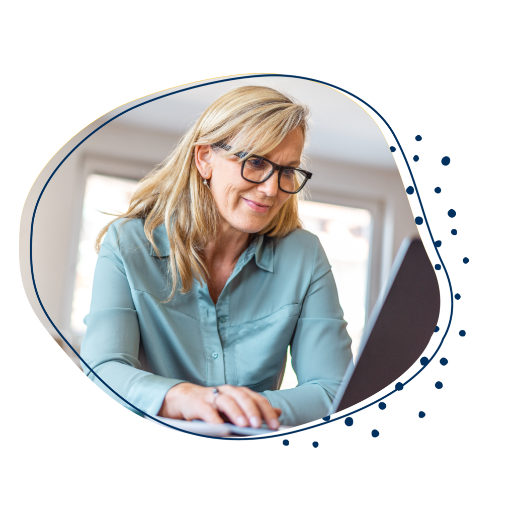 Ältere blonde Frau mit blauer Bluse sitzt am Laptop und liest auf diesem.