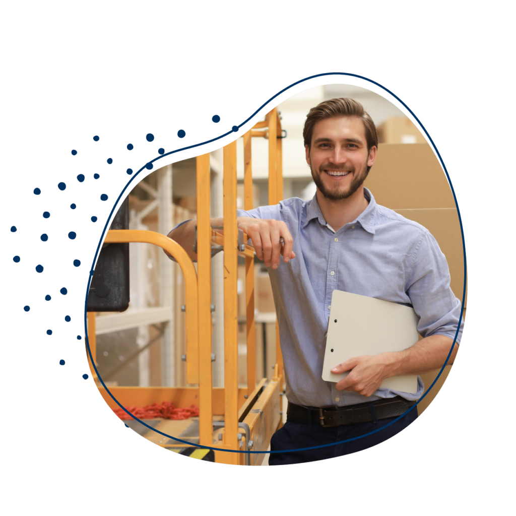 Ein lächelnder Mann mit Klemmbrett lehnt an einer orangenen Hebebühne in einem Lagerraum.