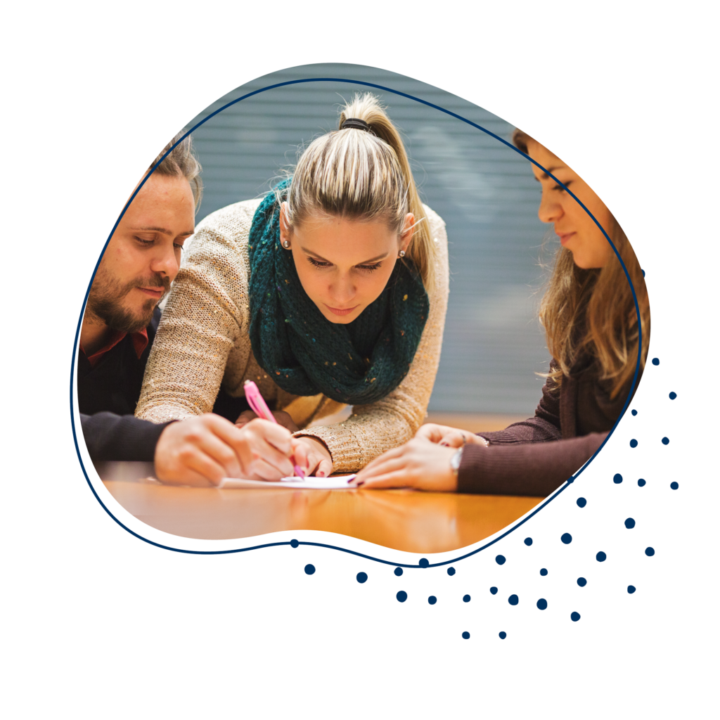 Drei Personen sitzen an einem Tisch und beugen sich über ein Blatt. Die Frau in der Mitte schreibt auf diesem.