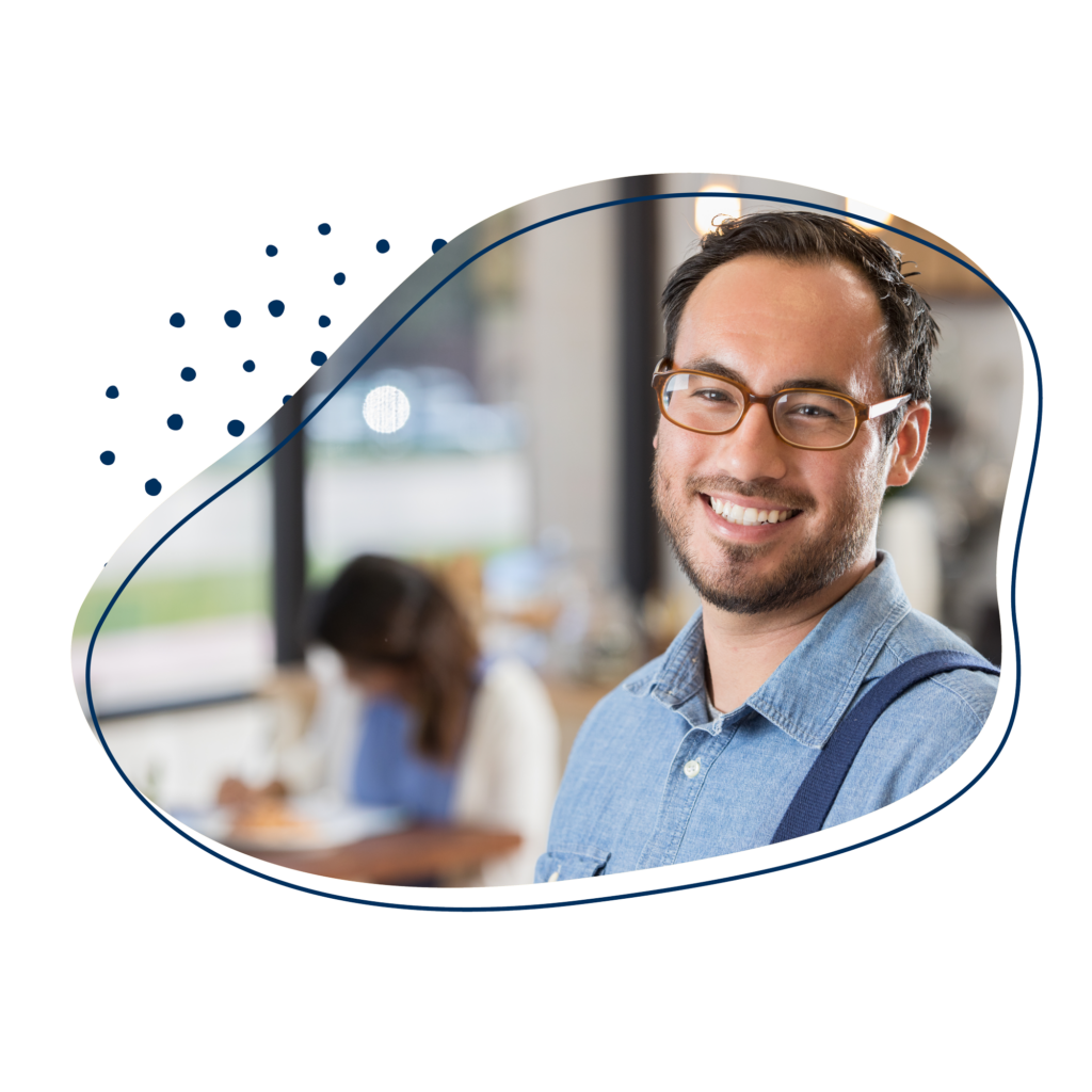 Mann im blauem Hemd lächelt in die Kamer, im Hintergrund ist eine Frau zu sehen, die an einem Schreibtisch arbeitet.