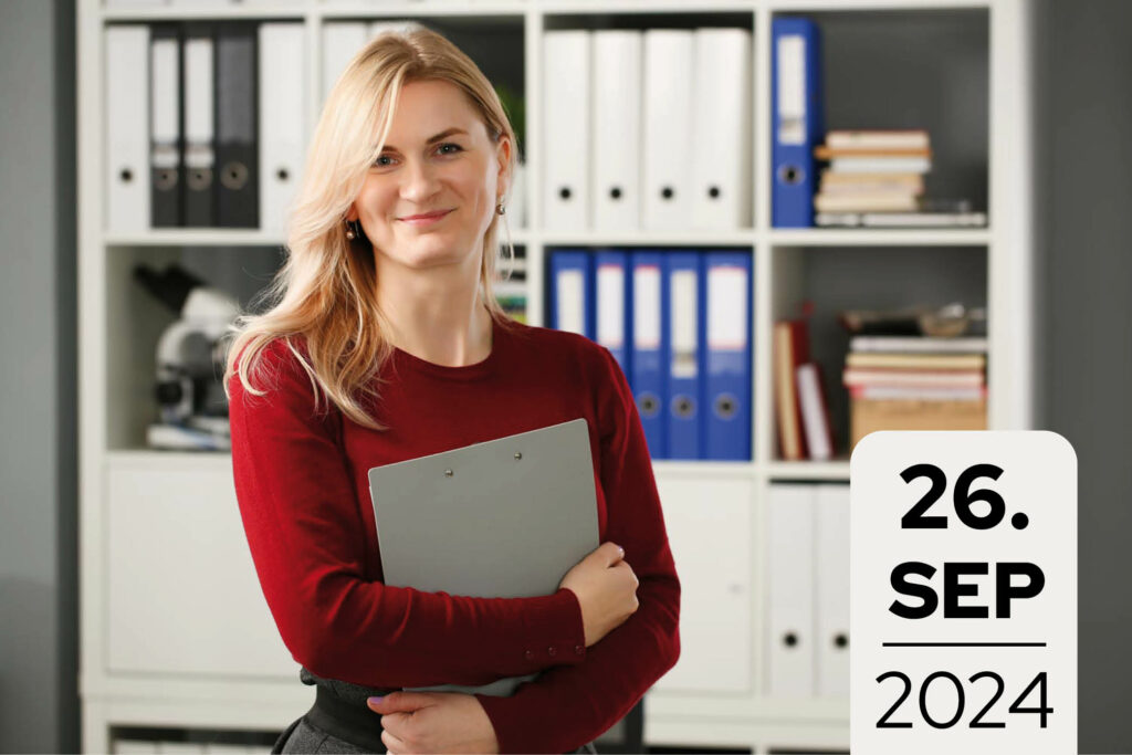 Junge Frau im roten mit Klemmmappe steht vor einem Regal an Aktenordnern