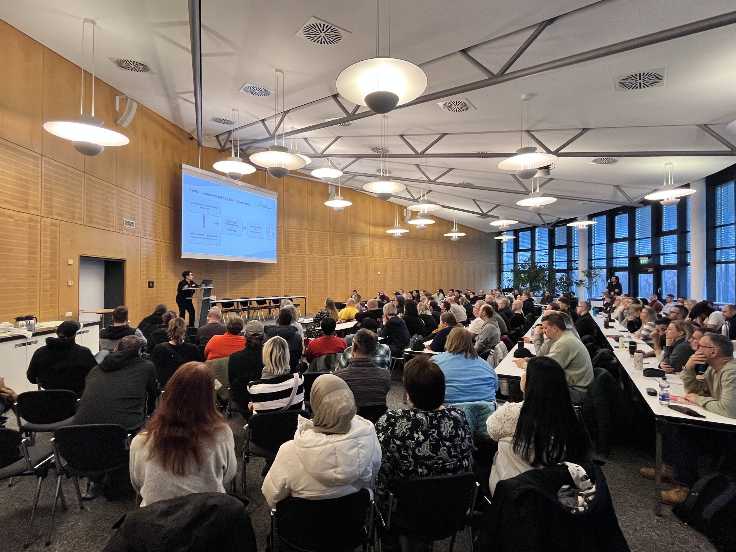Pädagogische Fachbereichsleitung begrüßt die neuen Umschüler:innen, die in Sitzreihen in der Aula des BFW Leipzig sitzen, zum Umschulungsstart.