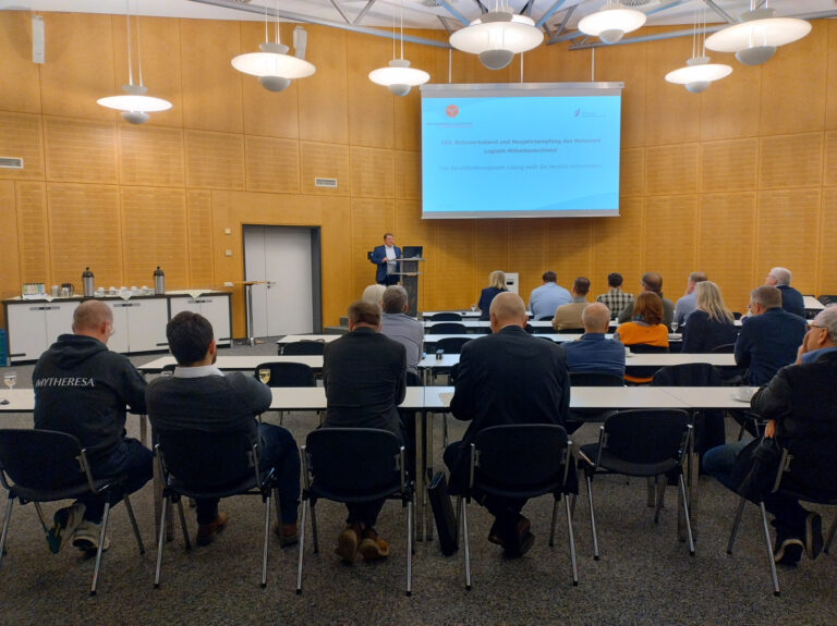 Foto: Eröffnung des Neujahrsempfangs des Netzwerk Logistik Mitteldeutschland in der holzvertäfelten Aula des BFW Leipzig. Zahlreiche Teilnehmende sitzen in Reihen auf Stühlen und hören aufmerksam zu.