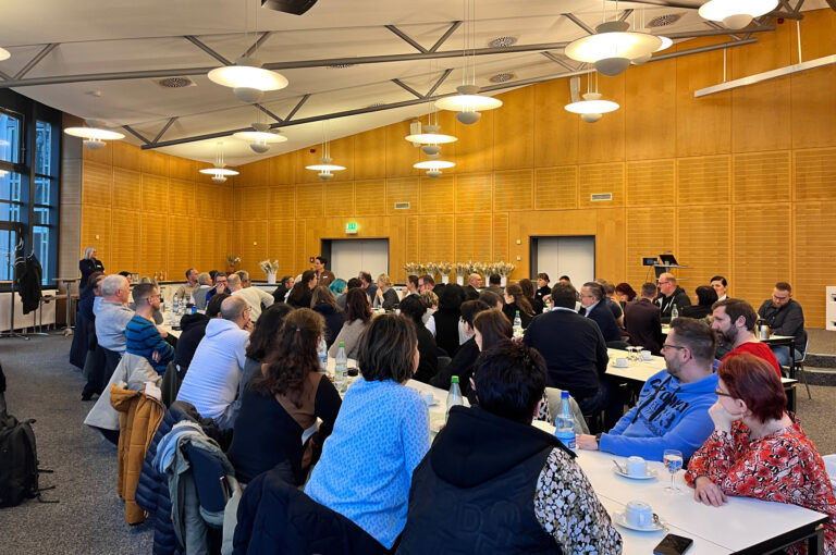 Absolventen sitzen an langen Tischen bei der Abschlussfeier ihrer Umschulung in der holzvertäfelten Aula des BFW Leipzig.