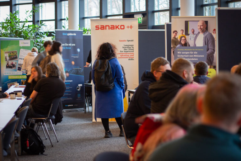 Mehrere Rollups der anwesenden Firmen auf dem Job-Buffet im BFW Leipzig stehen hintereinander. An den Ständen sitzen und stehen mehrere Menschen und unterhalten sich.
