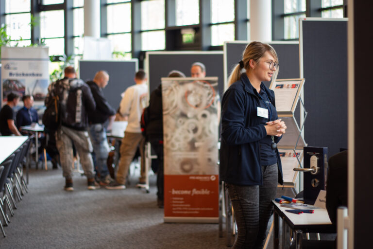 Blick in den Raum, in dem die Jobmesse im BFW Leipzig stattfand. Man sieht verschiedene Messestände. Im Vordergrund eine Personalerin an ihrem Messestand. Im Hintergrund sind unscharf mehrere Personen an den weiteren Messeständen zu sehen.